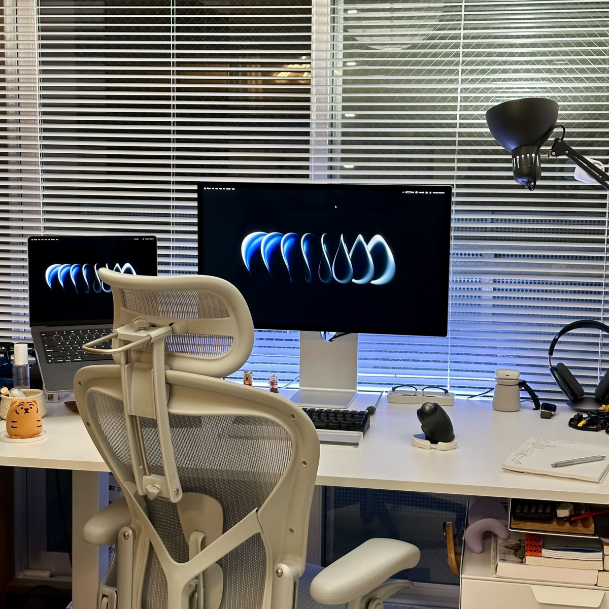 白いオフィスデスクの上に設置されたPCデスク環境。中央にはAppleのStudio Displayモニターと、その左隣にMacBookが置かれており、どちらの画面にも同じ青い抽象的なグラフィックの壁紙が表示されています。デスクの手前には、ヘッドレスト付きのグレーのアーロンチェアのような高機能メッシュワークチェアが後ろ向きに配置されています。デスク上には、トラックボールマウス、キーボード、ノートとペン、ヘッドホン、デスクライト、小さなトラの置物などが整然と並んでおり、背景の窓には白いブラインドが下がっています。
