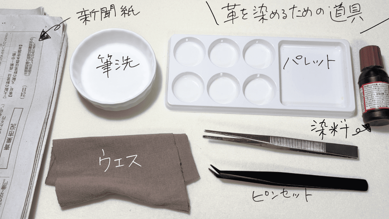 革を染めるための道具セット。新聞紙、筆洗、パレット、染料、ピンセット、ウエスが並んでいる。手書き文字で各道具の名前が記されている。