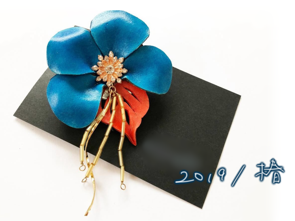 2019年に制作された椿の革花アクセサリー。光沢の強いつやあり仕上げが特徴で、中央には装飾パーツがつけられている。