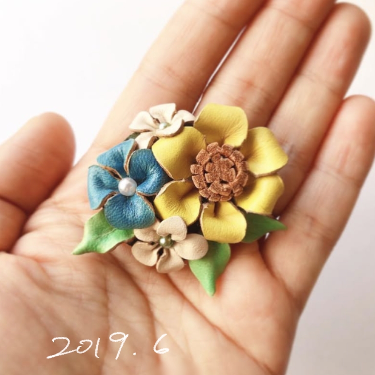 手のひらにのせた革花のブローチ。黄色・青・ベージュの花と葉が立体的に配置された作品（2019年6月制作）