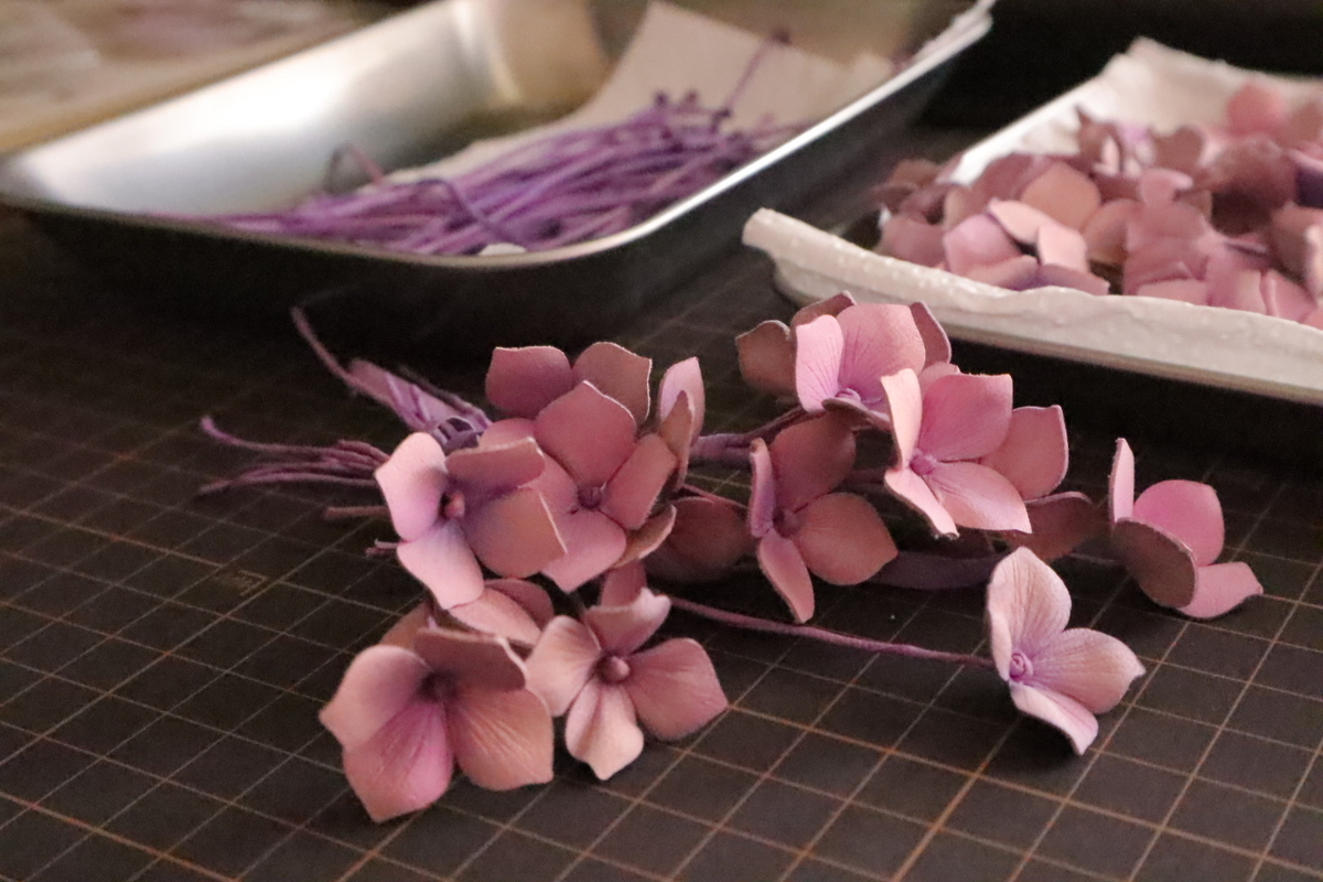 紫陽花ブローチの制作過程。細長く染められた茎がトレイに整えられ、手前には組み立て途中の紫陽花パーツが並んでいる。淡い紫とピンクが混ざり合い、繊細な色合いが特徴。
