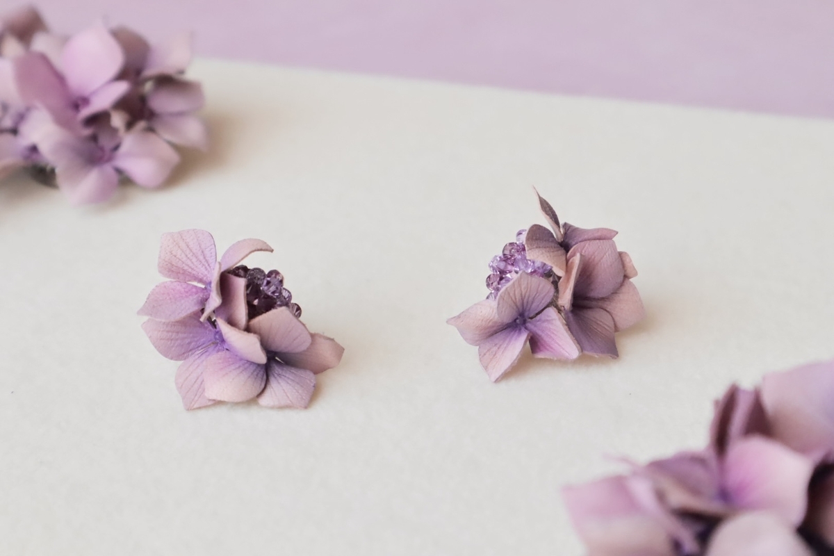 淡い紫色の革花ピアス。繊細な花びらと透明感のある紫のビーズが美しく配置されている。背景は白く柔らかな布で、周囲に同系色の革花がぼんやりと写っている。