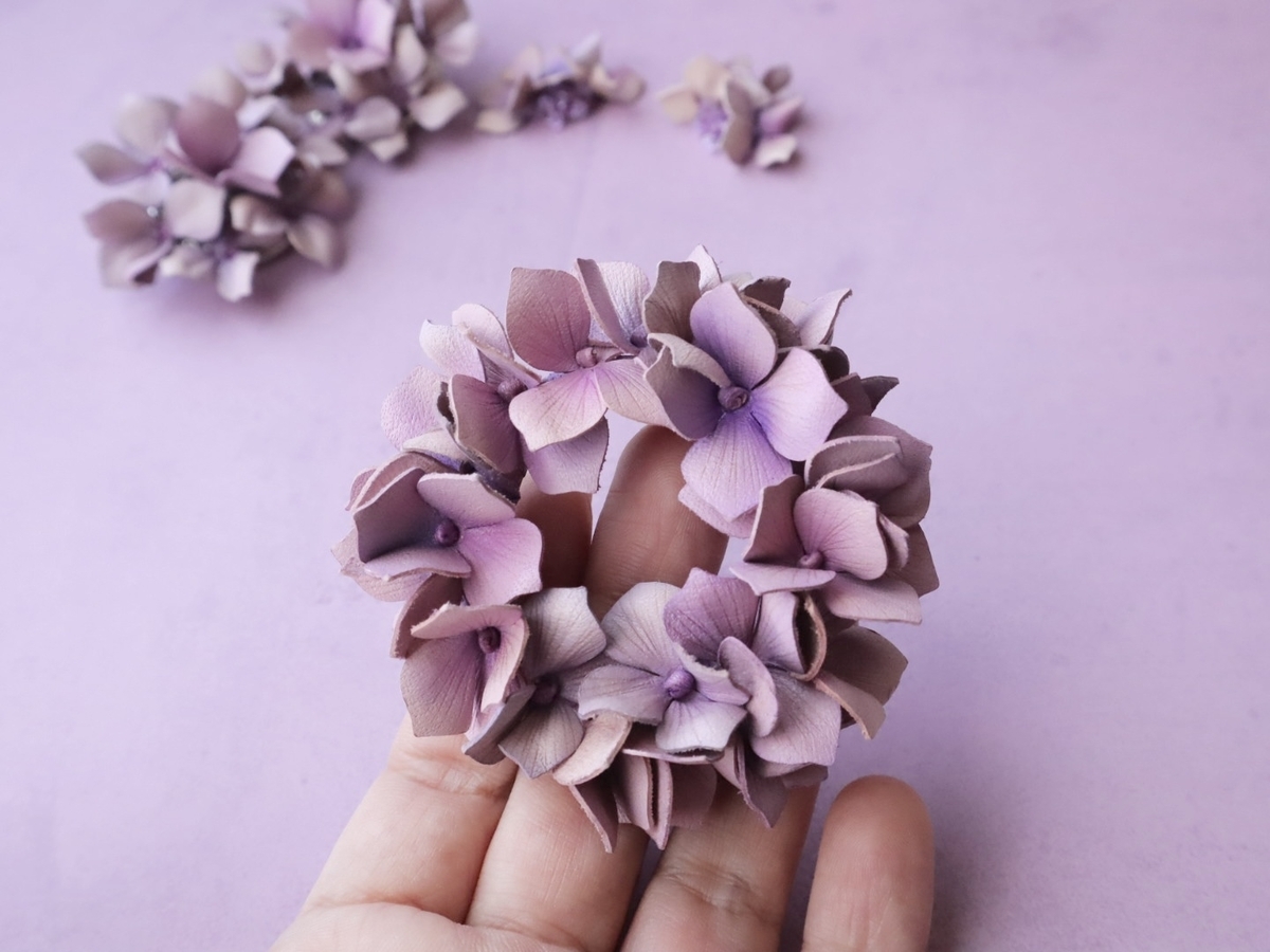 淡い紫色の革花アクセサリーを手に持つ様子。紫陽花の花びらが優しく重なり、名残惜しさの中に前向きな気持ちが重なる瞬間。