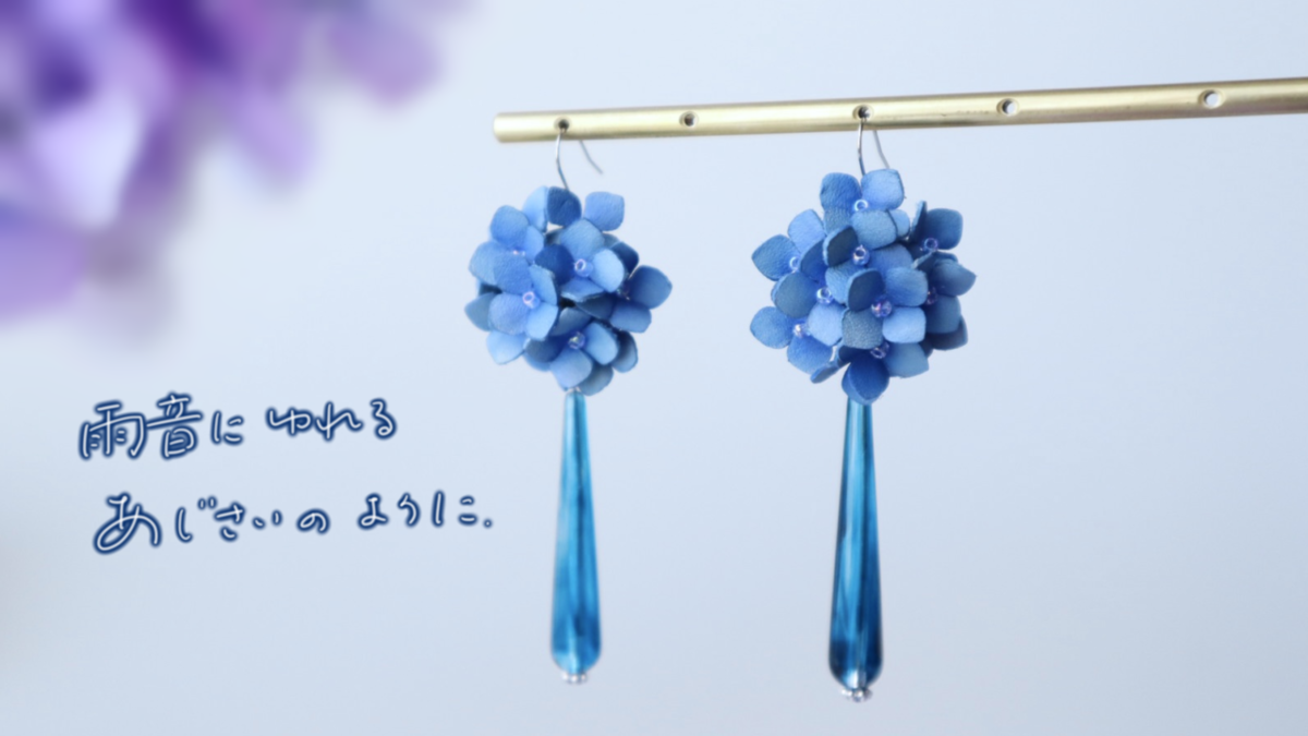 青い革の紫陽花をモチーフにしたピアスが2つ、しずく型のビーズとともに吊り下げられている。左下に「雨音にゆれる あじさいのように。」という詩的な文字が添えられている