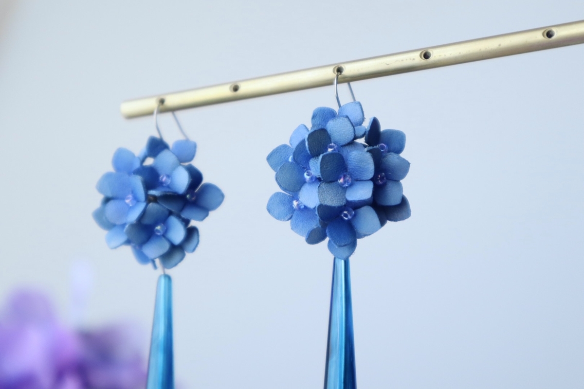 グラデーションが美しい青の革で作られた紫陽花モチーフのピアスが2つ、真鍮色のバーに吊り下げられている。紫陽花の花びらは立体的に重なり、中心に小さなビーズが輝いている。背景は淡いブルーで、左下にはぼかされた紫の花が見える。