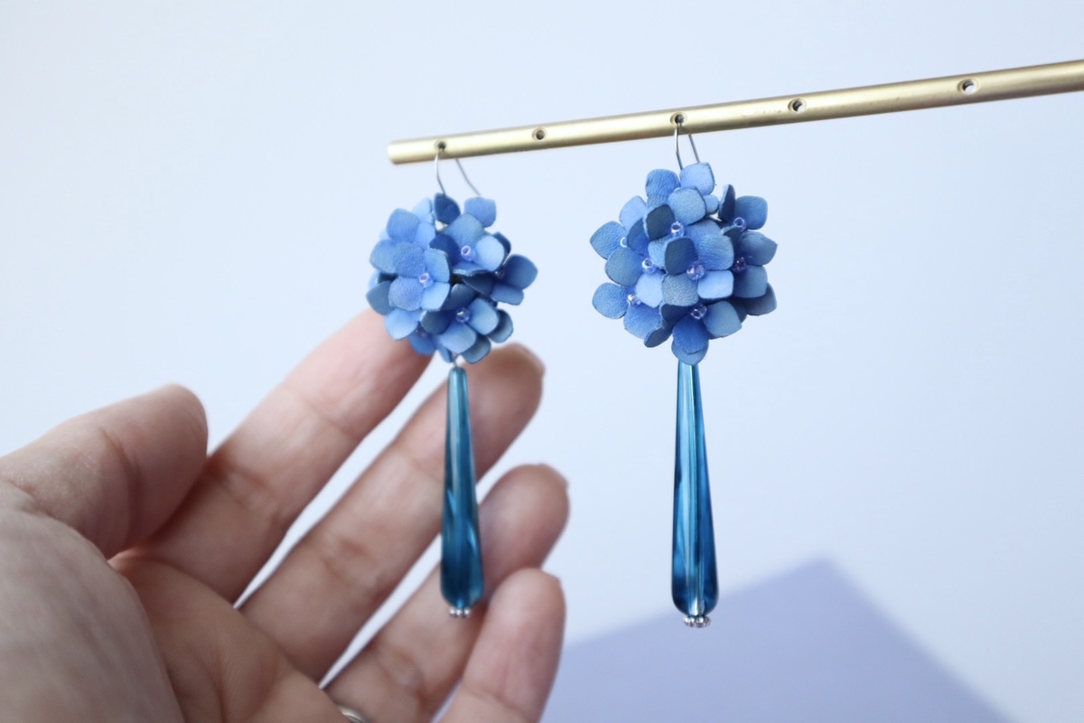 手のひらにかざされた青い革の紫陽花モチーフのピアスが2つ、真鍮色のバーに吊り下げられている。紫陽花は小さな革花を重ねて立体的に仕上げられ、下には青緑色のしずく型ビーズが揺れるデザイン。