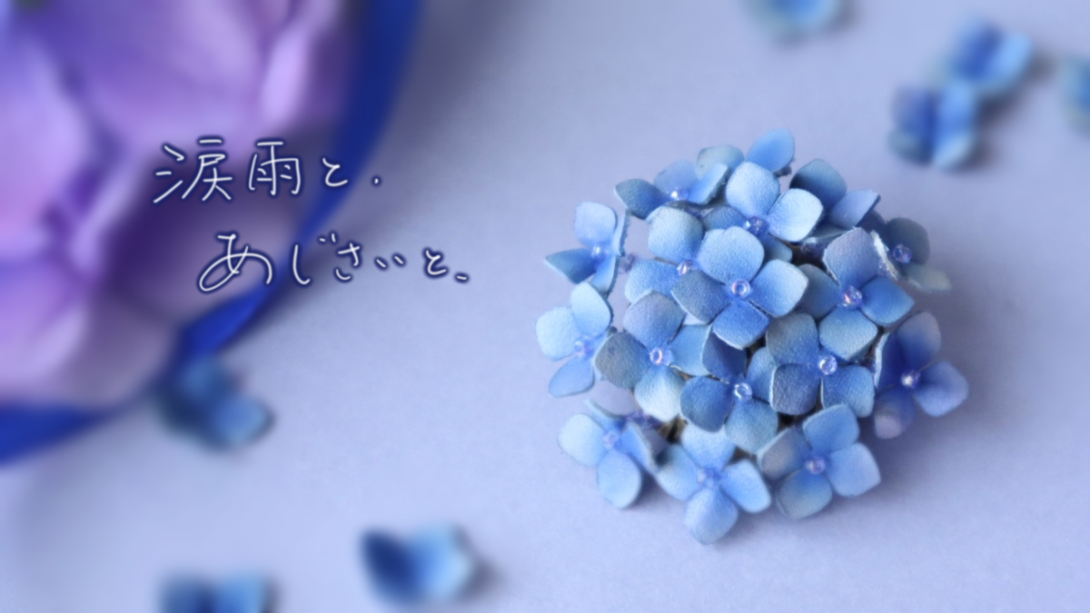青いグラデーションの革で作られた紫陽花のブローチが中央に置かれ、周囲には小さな花びらが散っている。左上には「涙雨と。あじさいと。」という詩的な文字が添えられ、背景には紫の紫陽花の花が大きくぼかされて映っている。
