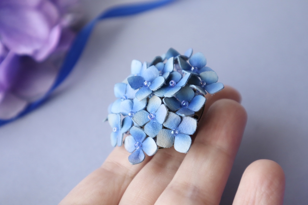 青い革で丁寧に作られた紫陽花モチーフのブローチが手のひらに乗せられている。花びらはグラデーション染めで仕上げられ、中心には小さなビーズが添えられている。背景にはぼんやりと紫の紫陽花と青いリボンが見える。