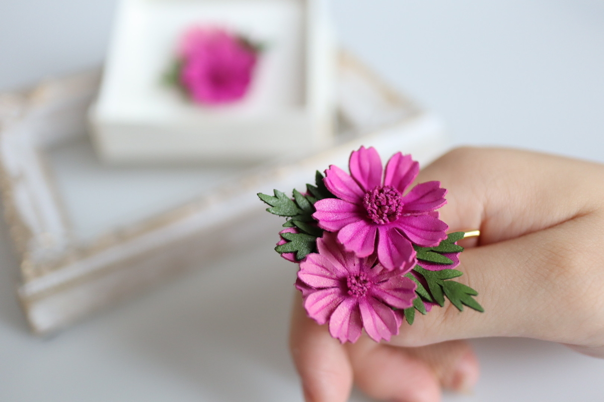 革で作られた秋桜のポニーフックを手に持った画像。ビビットカラーのピンク色の秋桜が大小二輪咲いており、鮮やかさが引き立つ。