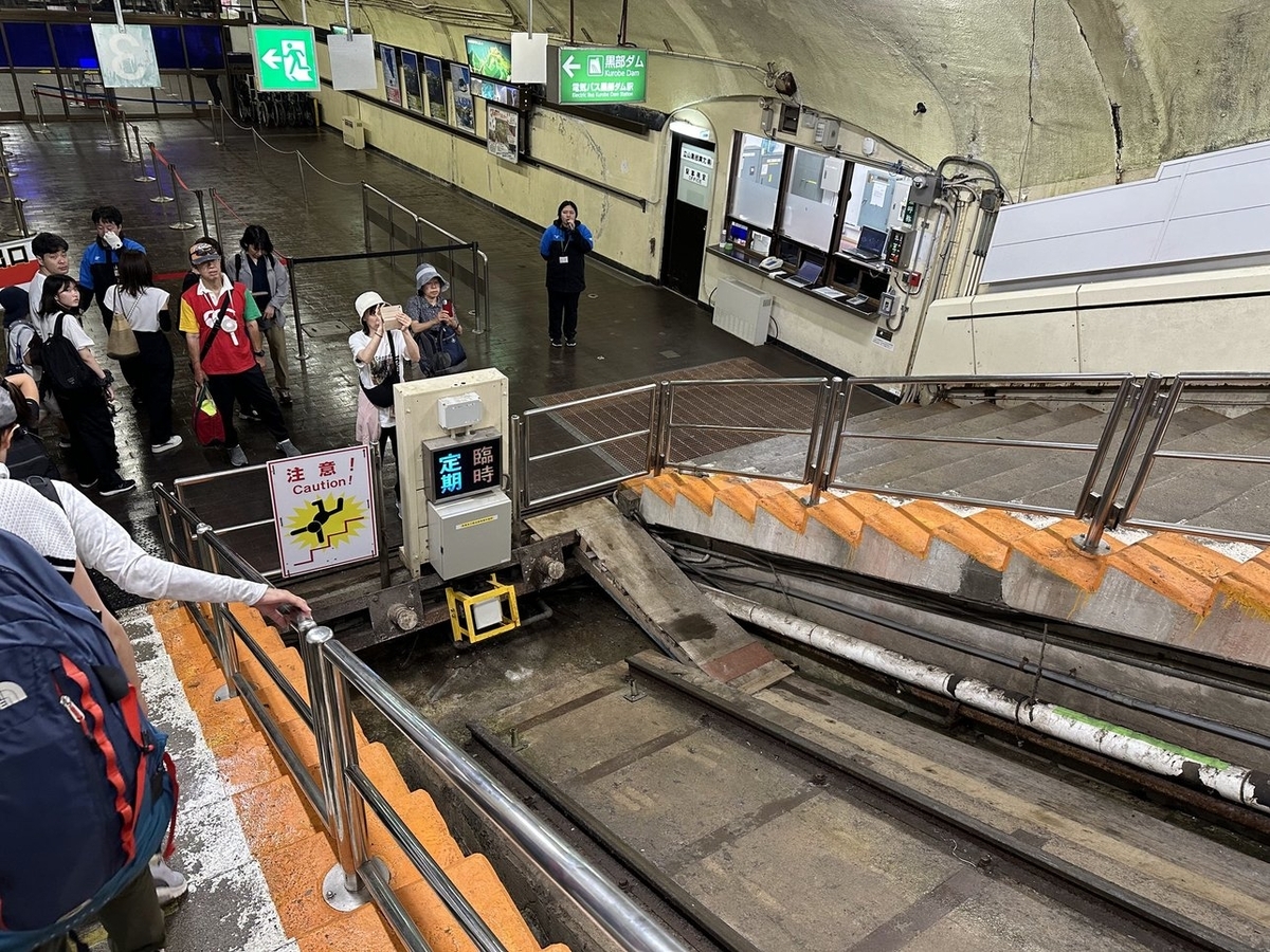 階段を降りる。奥に黒部ダムと書かれた案内板。ケーブルカーの線路が途切れた部分に「臨時 定期」と書かれた表示板があり、「定期」が点灯している。