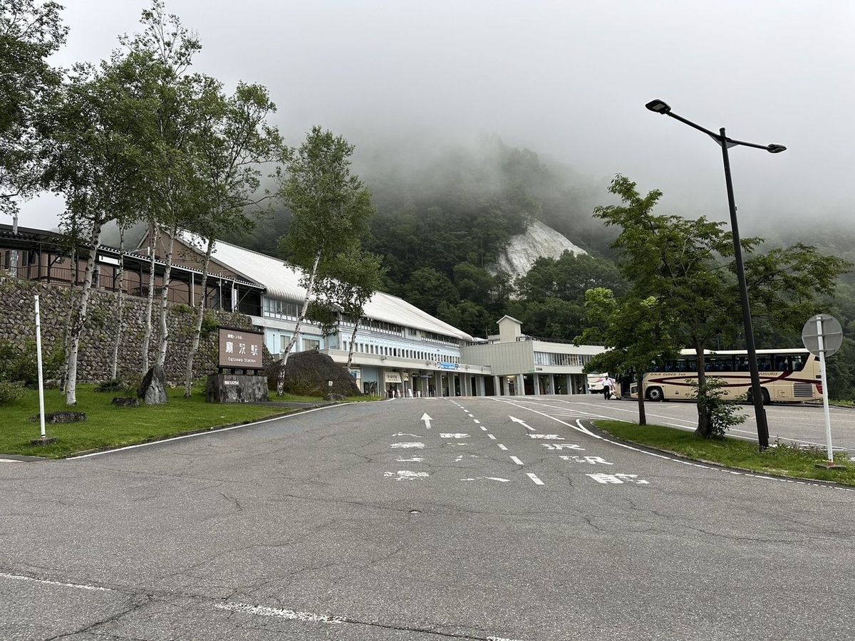 駅舎を左斜め前から撮影。中央に駅舎へ続く道路が、その左側に「関電トンネル 扇沢駅」と書かれた看板がある。駅舎は三角屋根で水色に塗装されている。