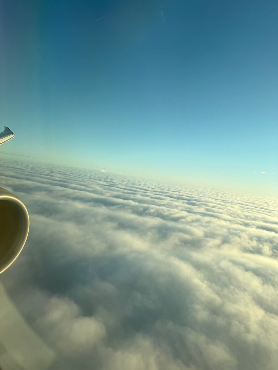 飛行機の窓から見える一面の雲海と青空。雲の上に広がる空は澄み渡り、太陽の光が雲に柔らかなグラデーションを作り出している。