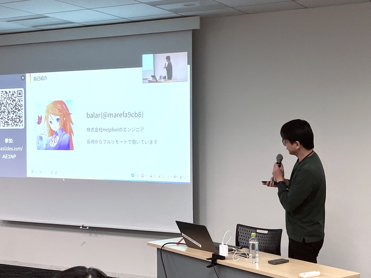 スライドがスクリーンに投影されており、その右側に男性がマイクを持って立っている