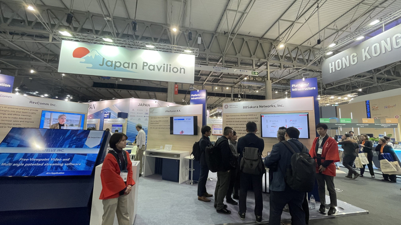 MWC2025展示会場内におけるJAPAN Pavilionを入り口正面から撮った画像。右側にBBSakura Networksのブースと、赤いハッピを来たBBSakura Networks社員がブース来場者を迎えている姿を映し出しています。