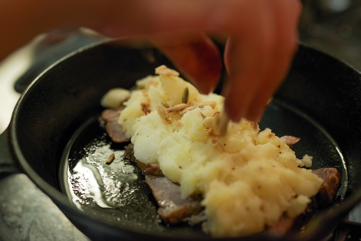 熱したスキレットに豚ローススモークとマッシュポテト、フライドオニオンとナッツ、その上からさらにスモークを