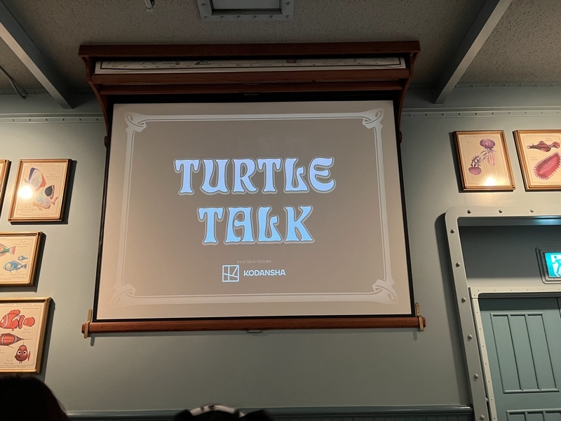 室内のプロジェクターに「TURTLE TALK presented by KODANSHA」と表示されている