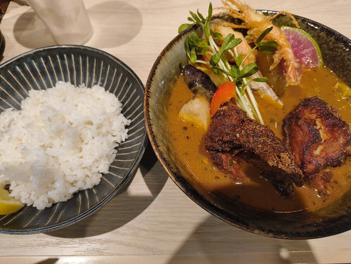 スープカレーの写真。左側にライスのお椀。右側にザンギ、水菜、甘エビの天ぷら、なすなどが入ったスープカレー。