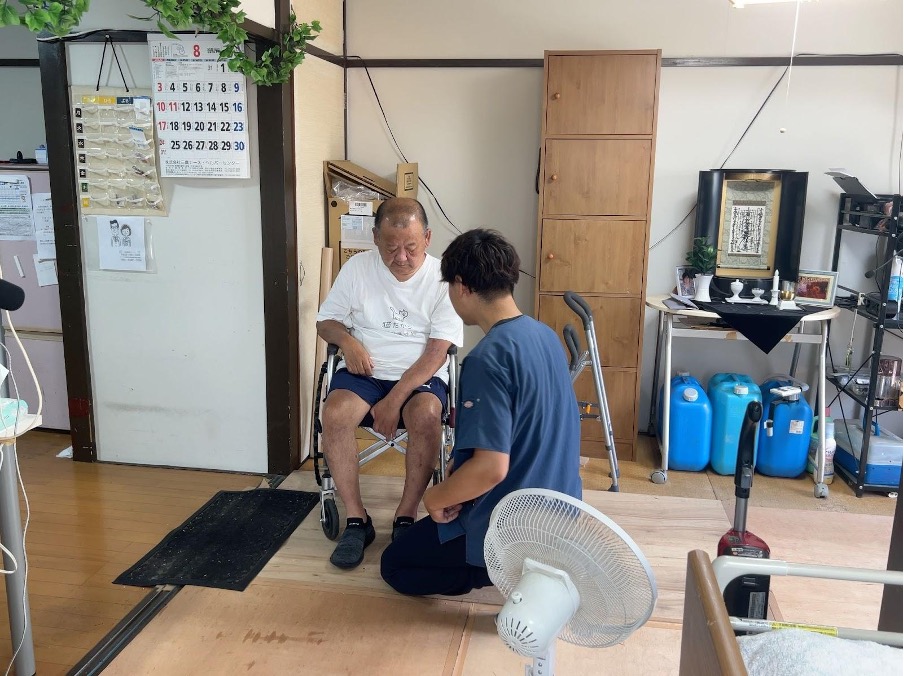 訪問リハビリを受ける男性と、セルフストレッチの方法を紹介するリハビリスタッフ