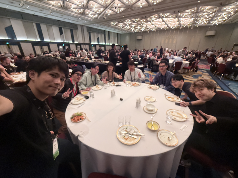 丸い机の周りに弊社のメンバーが並んで座り食事をしていて、カメラに向かってピースしている