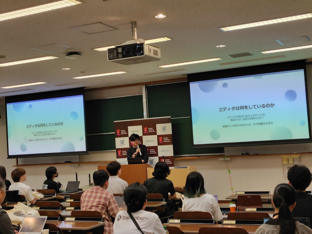 登壇中のコサキン。「エディタは何をしているのか」というスライドが左右のディスプレイに表示されている