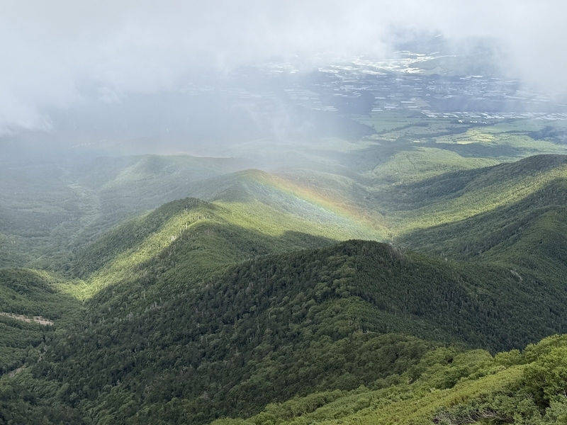 県界尾根から見下ろした樹林の尾根群にかかる虹と、その奥に広がる野辺山の高原野菜畑