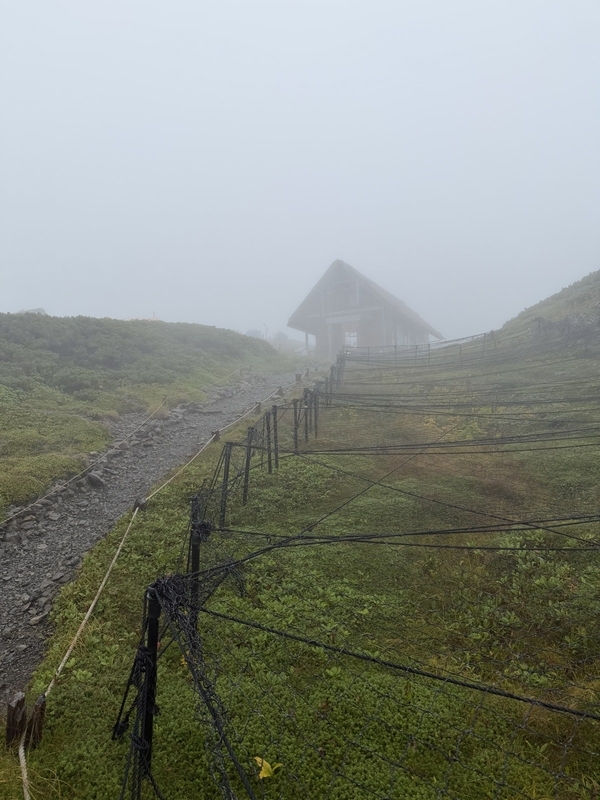ガスの向こうに荒川中岳避難小屋の屋根が見えてくる。手前には防護柵の続く登山道