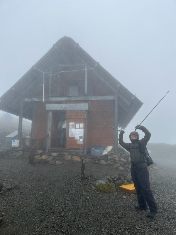 ガスに包まれた荒川中岳避難小屋の三角屋根と、その前に立つ登山者