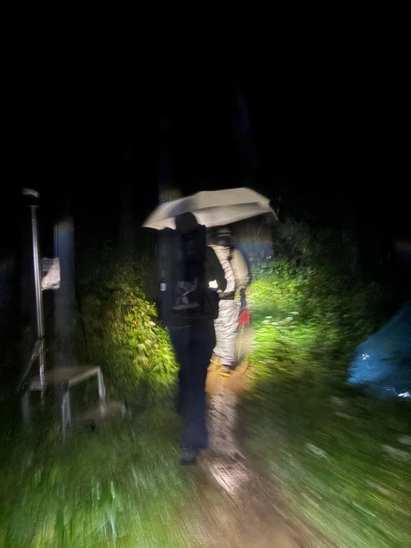 暗いテント場で、ヘッドランプの光に照らされた白い傘を差して歩く登山者