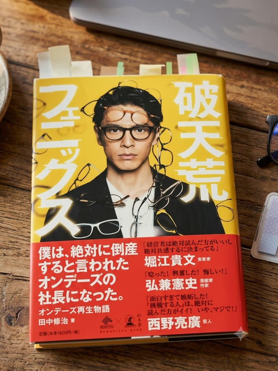 カフェ風の木目調テーブルに置かれた書籍『破天荒フェニックス オンデーズ再生物語』(田中修治 著)のクローズアップ写真。黄色の表紙には、複数のメガネをかけた著者のポートレートが描かれている。上部には複数の付箋が貼られ、下部の赤色の帯には「僕は、絶対に倒産すると言われたオンデーズの社長になった。」というコピーと、堀江貴文氏、弘兼憲史氏、西野亮廣氏による推薦コメントが掲載されている。背景には、わずかにノートパソコンとメガネのフレームが映り込んでいる。