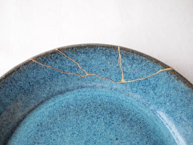 ​割れた青い陶磁器のヒビを金で美しく修復した「金継ぎ」の皿のアップ。失敗から学びリカバリーすることで価値が高まるキャリアのメタファー。