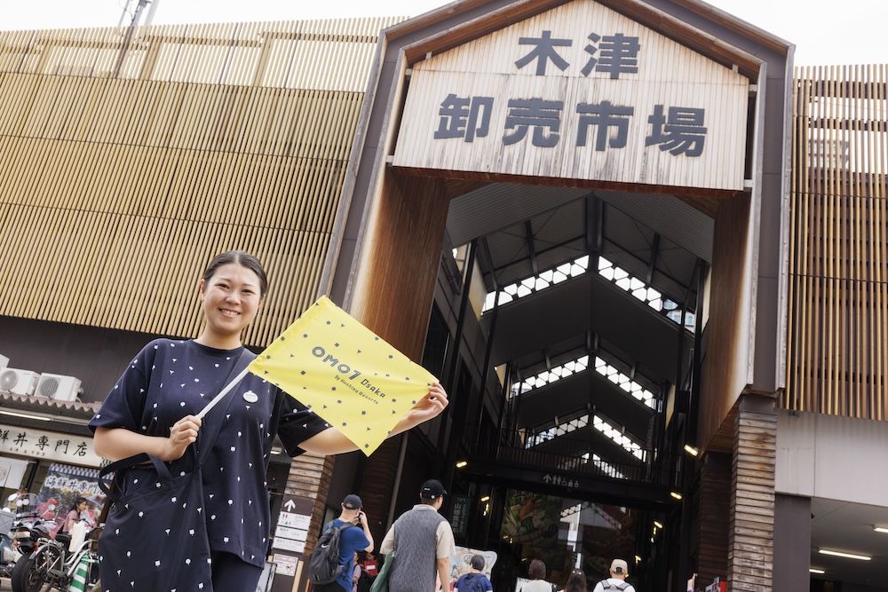 OMO7の旗をもって木津卸売市場の前に立つ女性