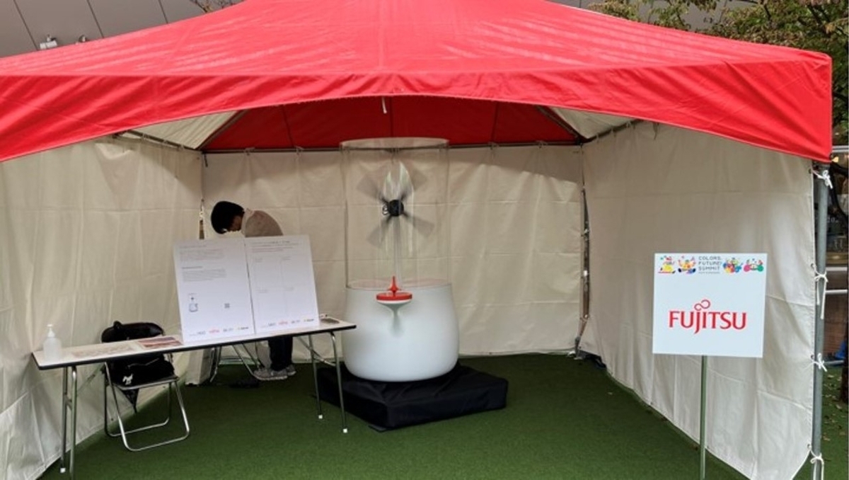 Colors, Future! Summit 2024に出展した際のラゾーナ川崎のルーファ広場の展示ブースの画像。緑の芝生の上に赤色の屋根のテントが置かれている。そのテントの下にワンダリングアーカイブスの筐体に加えて、本取り組みの説明パネルや富士通のロゴパネルも配置されている。