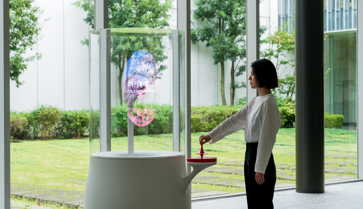 ワンダリングアーカイブスの動作イメージを表した画像。オレンジ色のコマを回すことで、白い台の上にのったプロペラ型のディスプレイに「未来のまちの画像」と「未来のまちで暮らす人の物語」が投影される。