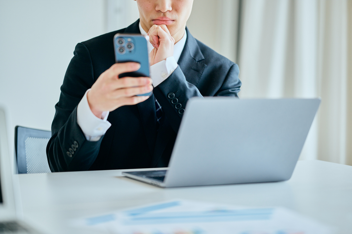 パソコン前でスマホをもって悩む男性