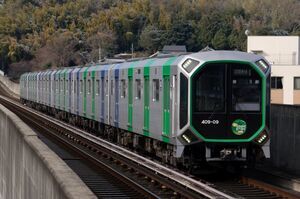 学研北生駒駅付近のストレートの高架線を走るOsakaMetro400系(406-09編成・「夢洲2025」HM付き)。宇宙船のような見た目の未来的なデザインで、テーマカラーは緑。