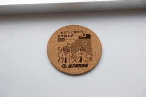 コルク製の丸形装飾。「あなたと越えたい赤字越え」と書かれている。イラストは歌う猫と収益グラフ