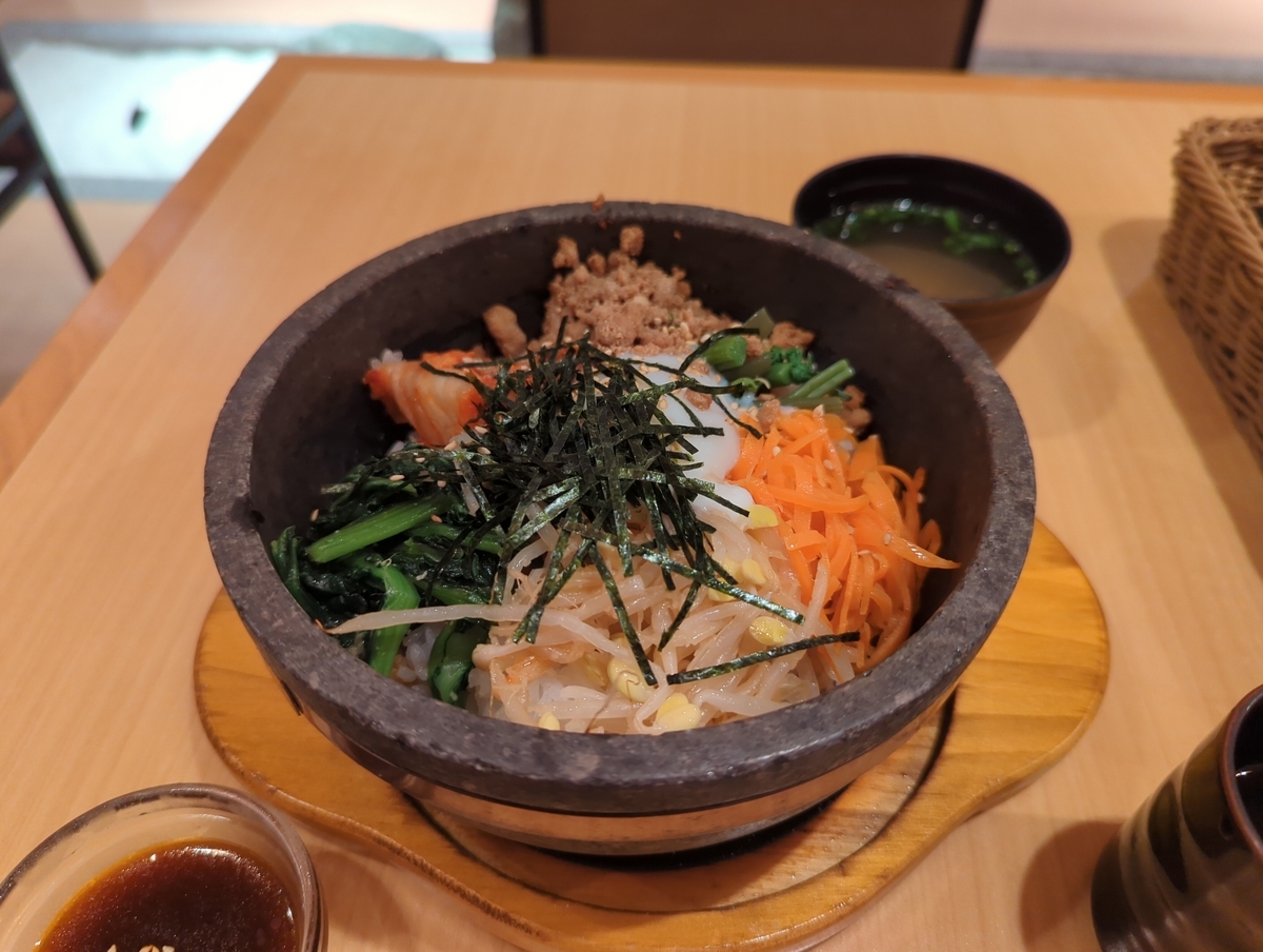 机の上に置かれた石焼きビビンパを写した写真．丼のごはんに各種のナムル，キムチ，温泉たまご，肉のそぼろ，刻み海苔などが乗せられている．ビビンパの周りには湯呑や汁物，コチュジャンの入った器が見える．