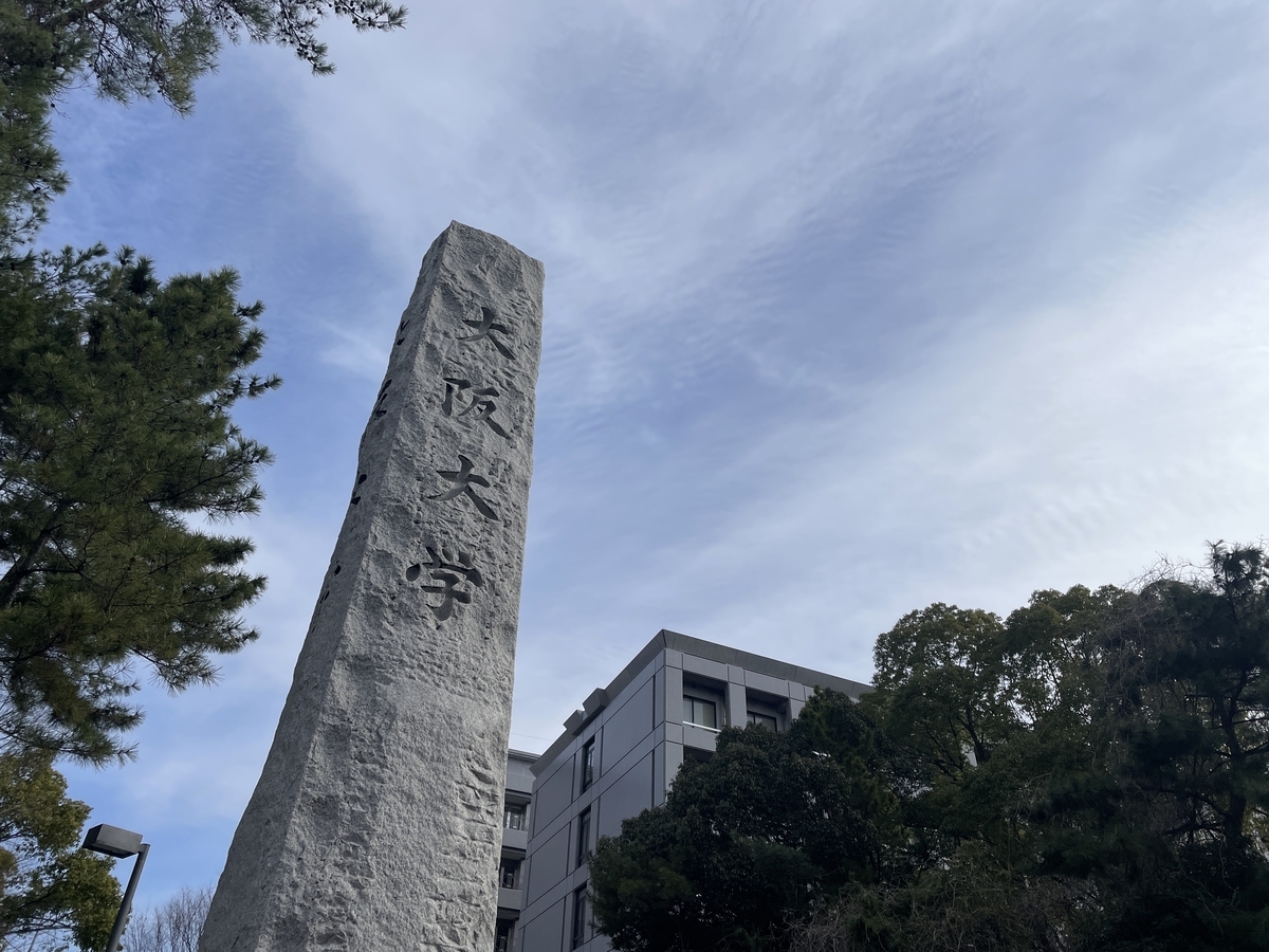大阪大学豊中キャンパスの正門の写真です。「大阪大学」と掘ってある石の塔がズドーンと立っています