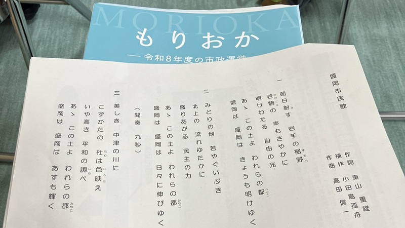 「盛岡市民歌」の歌詞が印刷された紙と、市政運営方針の冊子が机の上に置かれている。地区まちづくり懇談会の冒頭で市民歌斉唱が行われた。