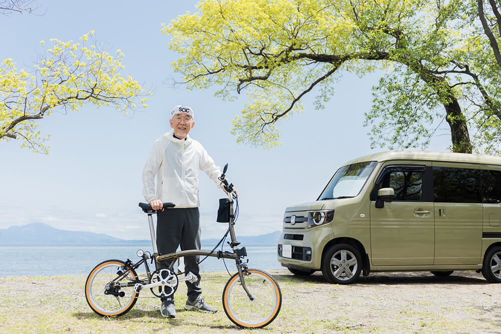 森の中のキャンプ場にて、N-VANの横で自転車を支えながら立つYouTuberのウィンピージジイさん