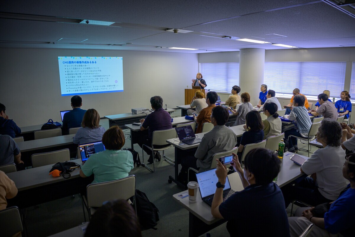 会議室で行われているセミナーの様子。山本和泉が前方でマイクを持ってプレゼンを行い、スクリーンには『CMS運用の画像作成あるある』と書かれたスライドが映されている。参加者たちはノートパソコンやタブレットを使いながら熱心に話を聞いている。