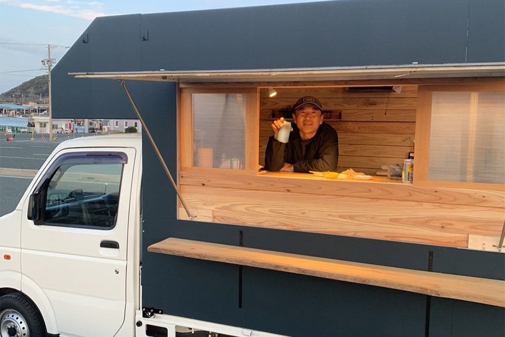 お手製のキッチンカーのカウンター越しにドリンクを飲むAtsuyaさんのお父さん