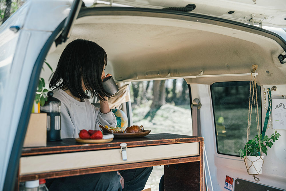 エブリイ車内に設えたベッドの上に腰かけながらコーヒーを飲むいとうさん