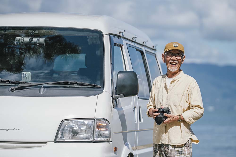 琵琶湖をバックにボンゴ横でカメラを手に持つウィンピーさん