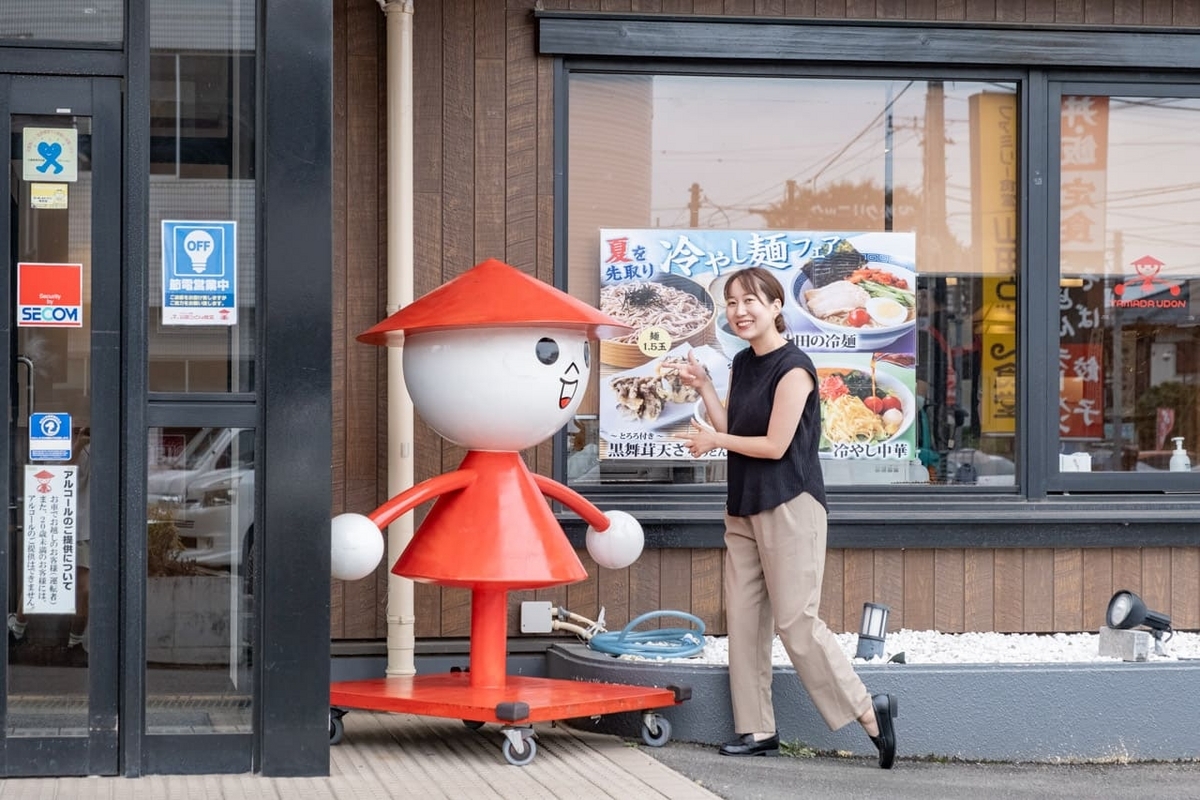 山田うどん食堂１号店の入り口でかかしくんと記念撮影する山口真央さん