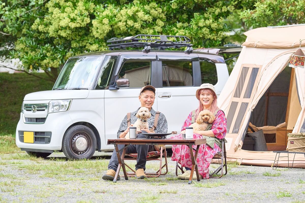 N-BOXとテントの前で椅子に座り、トイプードルを膝に乗せている、笑顔のさいとう夫婦