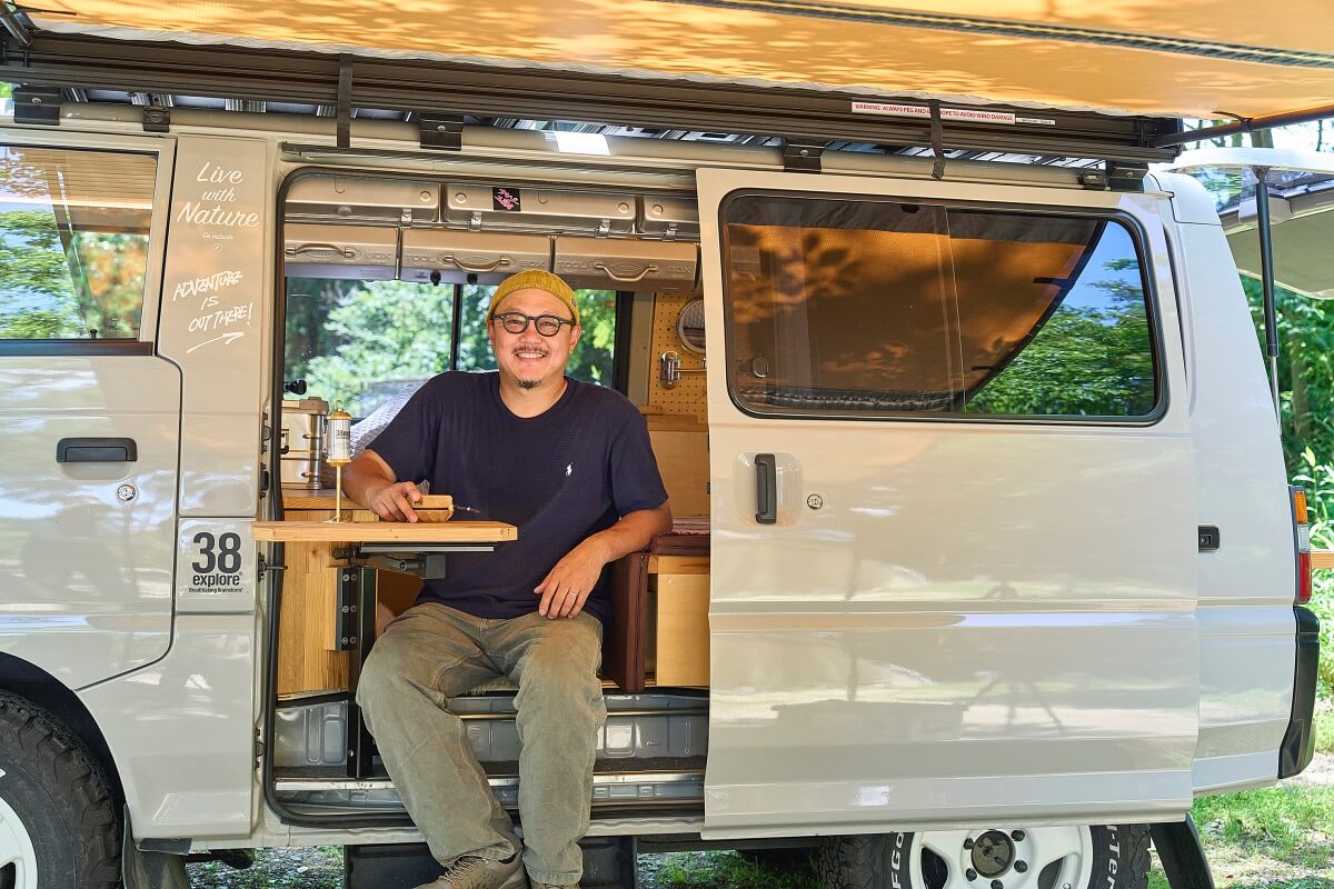 三菱デリカを車中泊カスタム。中古のデリカをキャンピングカーに改造。車中泊カスタムしたデリカバンのサイドドアを開けて座り微笑む宮﨑秀仁さん