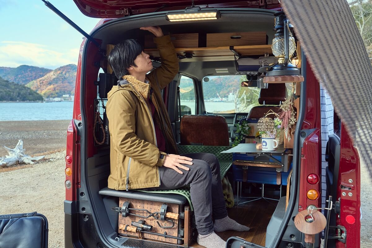 N-VANデナリの車内でベッドに腰掛け天井に手を伸ばすジベキャンさん