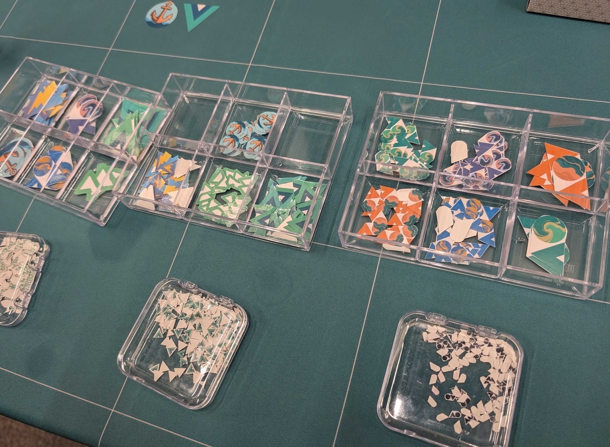 緑系のテーブルクロスの上に、透明の仕切り付きケースが並び、Vue や Vite などのロゴステッカーが仕分けられている。