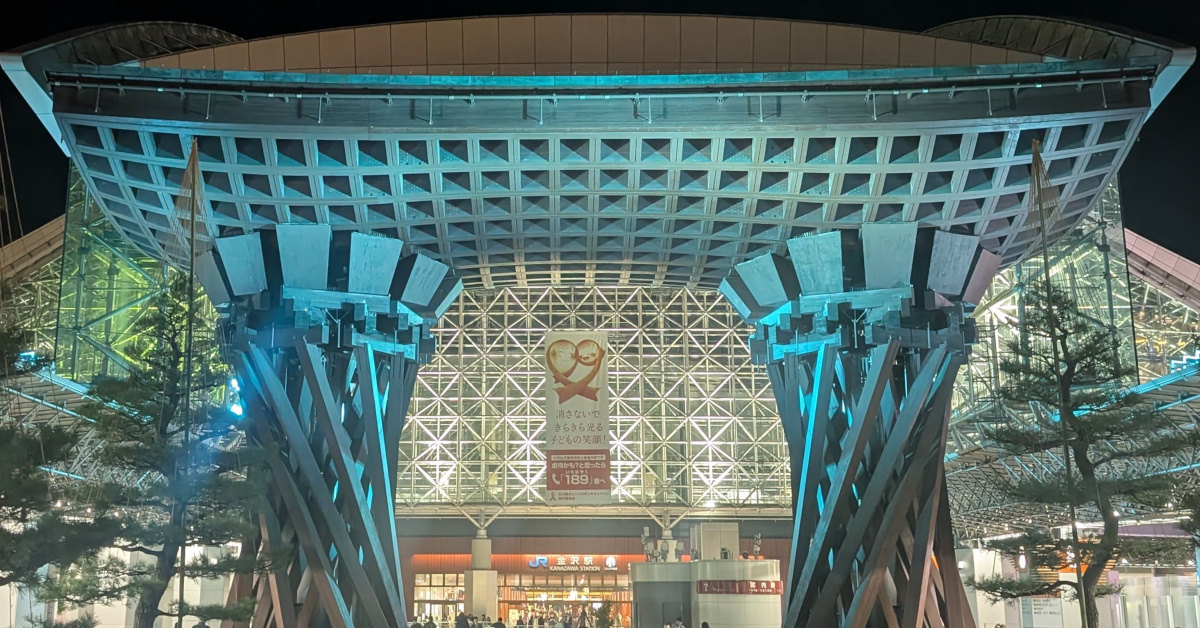 夜の金沢駅の遠景。鼓門を正面に据え、青緑色のライトアップがされている。奥のガラス張りの構内に垂れ幕と駅名看板が見える。
